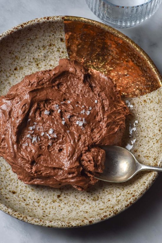 An aerial image of a beige ceramic speckled bowl filled with 3 ingredient chocolate mousse. The bowl sits on a white marble table and a glass of water sits in the top right corner.