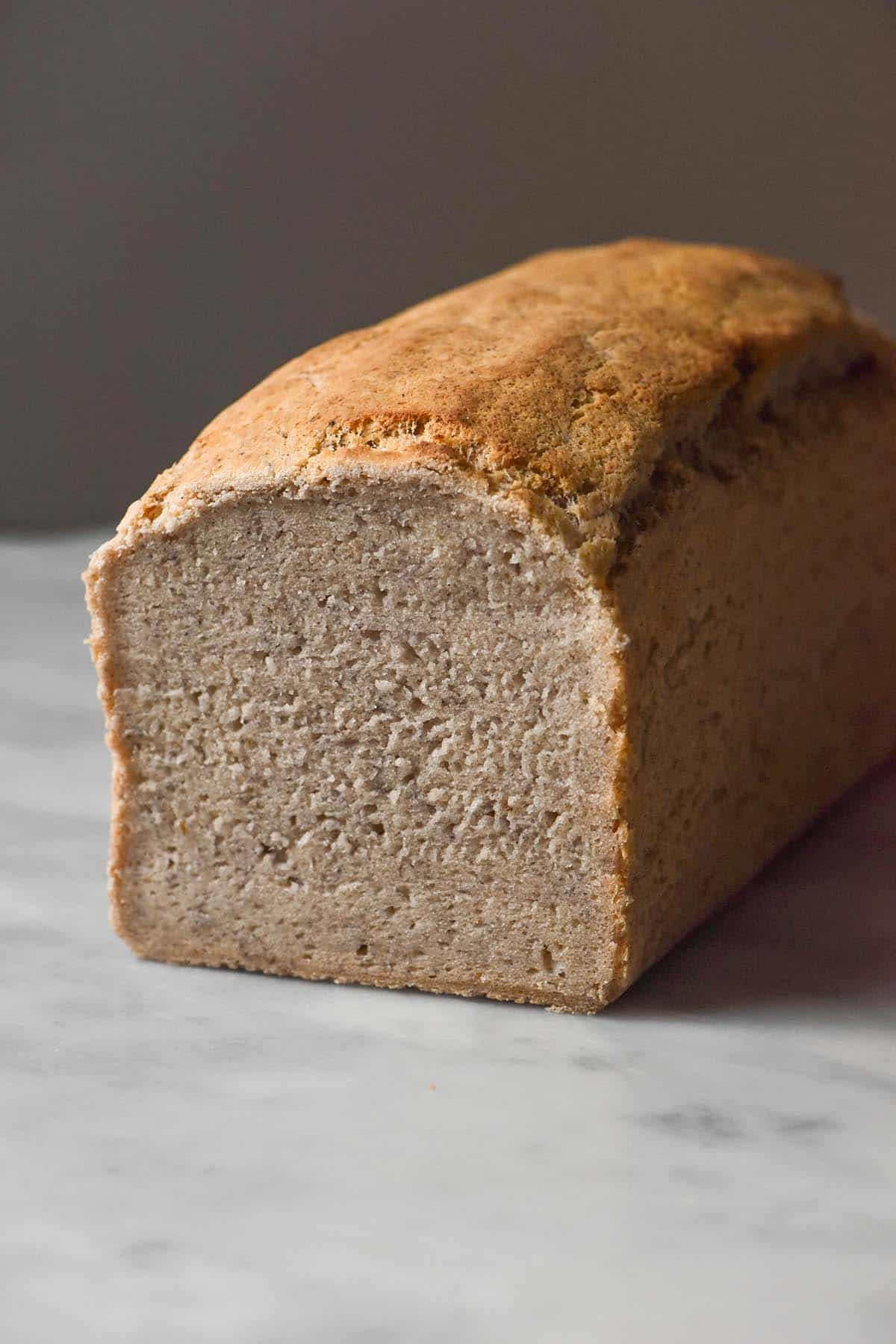 A side on image of a loaf of gluten free low histamine bread on a white marble table against a dark backdrop. The bread has been sliced revealing the crumb inside. 