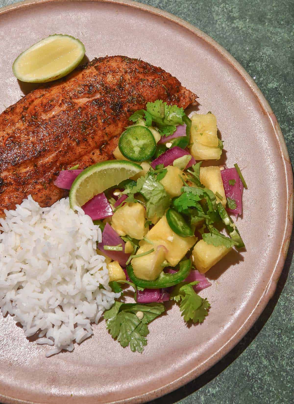 An aerial image of low FODMAP jerk fish, pineapple salsa and rice on a pale pink ceramic plate atop a dark olive backdrop