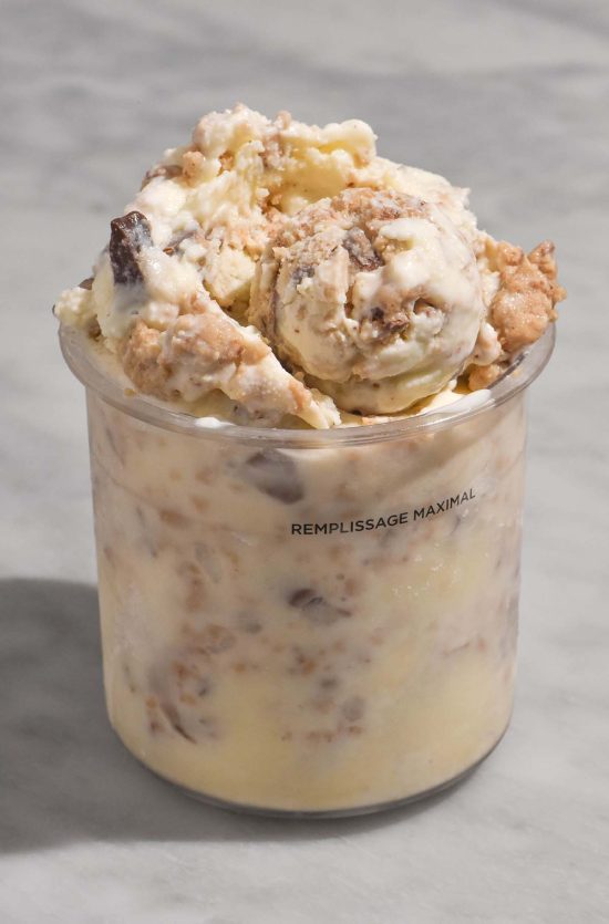 A side on image of a tub of Ninja Creami cookie dough ice cream on a white marble table. The tub has a scoop nestled in the top of that is filled with choc-chip cookie dough