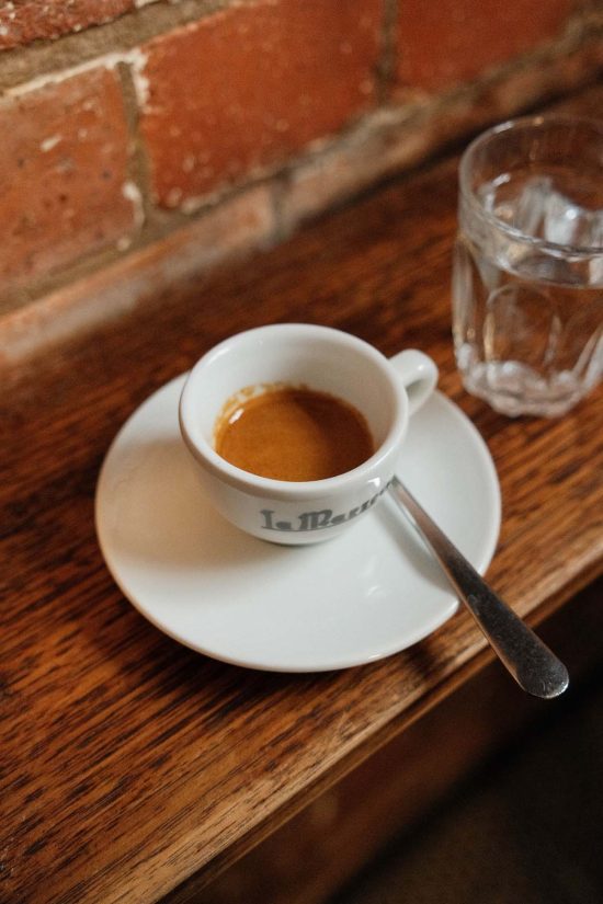 An image of a white espresso cup filled with espresso on a white saucer atop a wooden bench top against a brick wall. An espresso from John Mills Himself in Brisbane CBD.