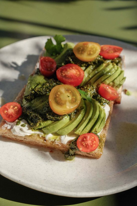 An aerial sunlight view of a gluten free avocado, pesto and tomato focaccia from Vanilla Foods in Noosa, Queensland. The plate sits on a dappled olive green table