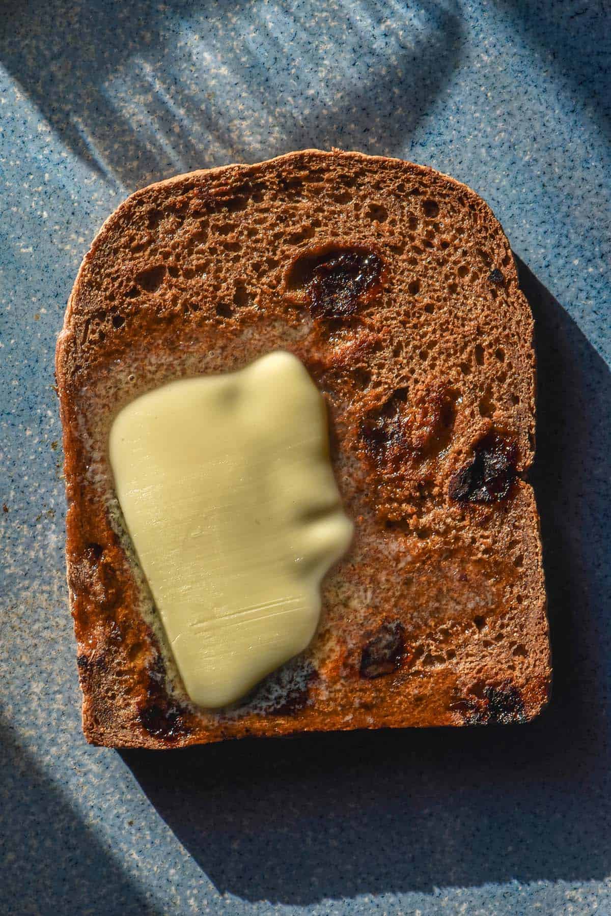 An aerial image of a slice of gluten free cinnamon raisin toast topped with butter. The toast sits on a bright blue ceramic plate in contrasting sunlight