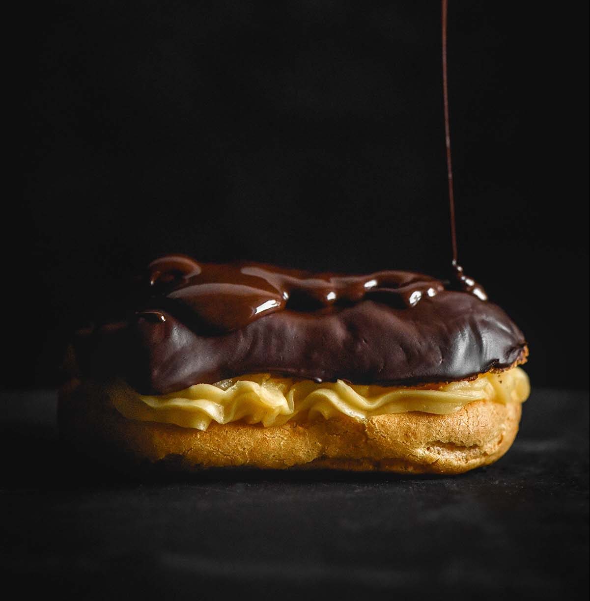 A dark and moody side on image of a gluten free eclair with chocolate drizzling down onto the top.