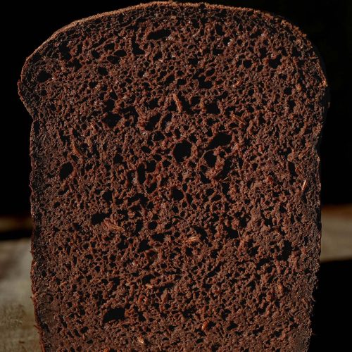 A macro close up image of a loaf of gluten free pumpernickel bread against a dark backdrop. A slice has been cut off revealing the soft inner crumb