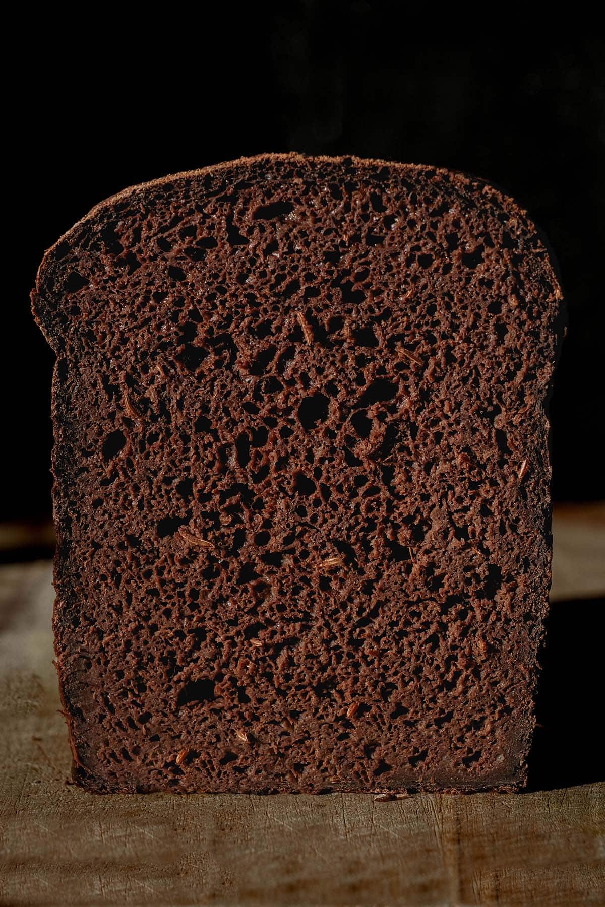 A macro close up image of a loaf of gluten free pumpernickel bread against a dark backdrop. A slice has been cut off revealing the soft inner crumb