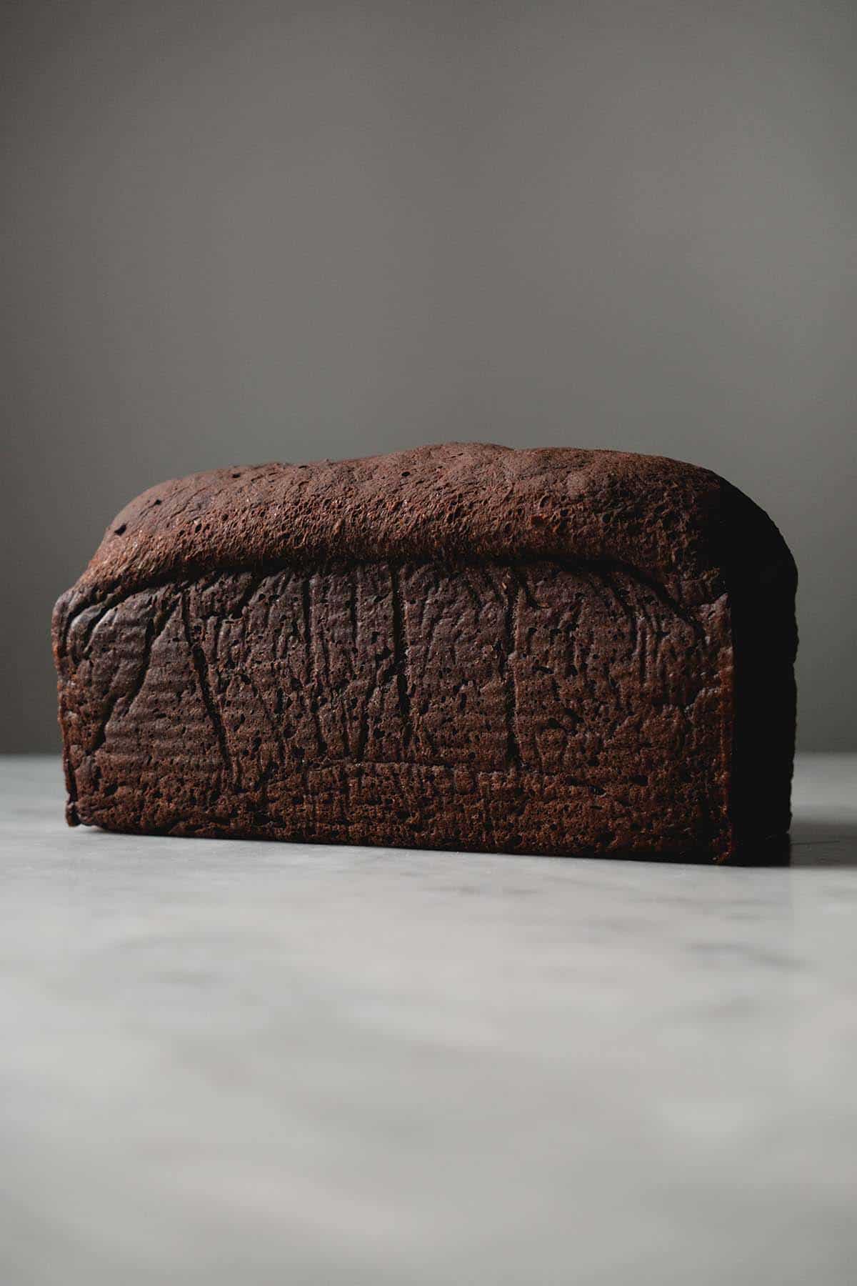 A side on view of a loaf of gluten free pumpernickel bread on a white marble table against a white backdrop