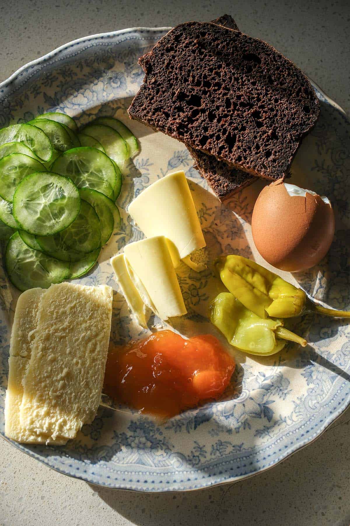 An aerial image of a Scandinavian style snack plate with gluten free pumpernickel bread. The food sits atop a white and blue patterned plate atop a stone bench top
