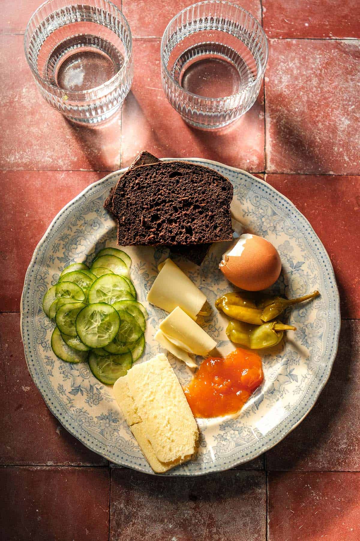 An aerial image of a snack plate topped with gluten free pumpernickel bread and accoutrements. The place sits atop a terracotta tile backdrop and two water glasses sit in the top of the image. 