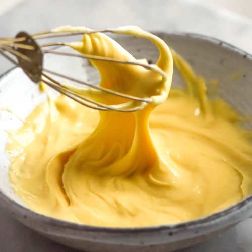 A bright and airy macro image of a bowl of lactose free pastry cream being whisked in a white bowl against a white backdrop