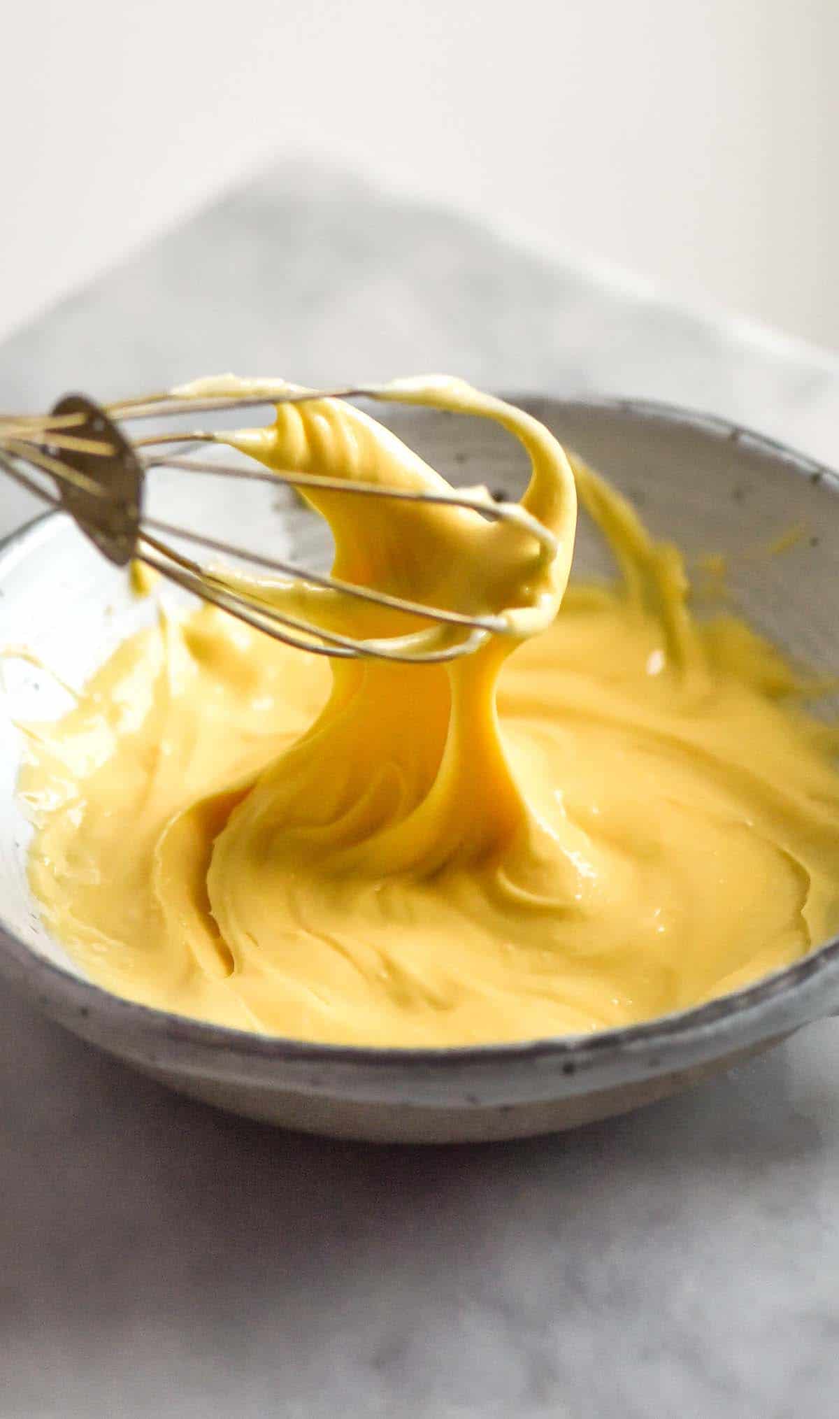 A bright and airy macro image of a bowl of lactose free pastry cream being whisked in a white bowl against a white backdrop