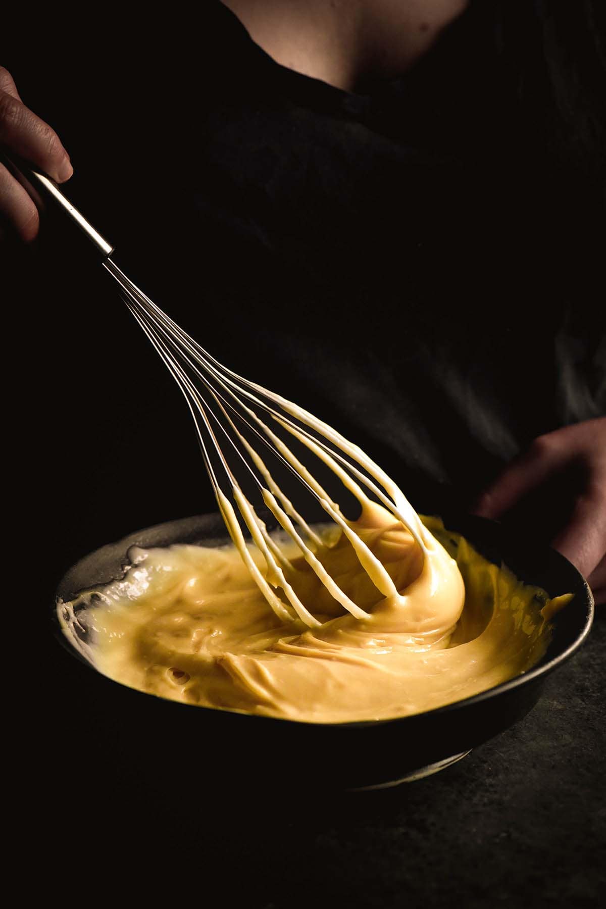 A moody image of a person in a dark shirt whisking a bowl of lactose free pastry cream against a dark backdrop