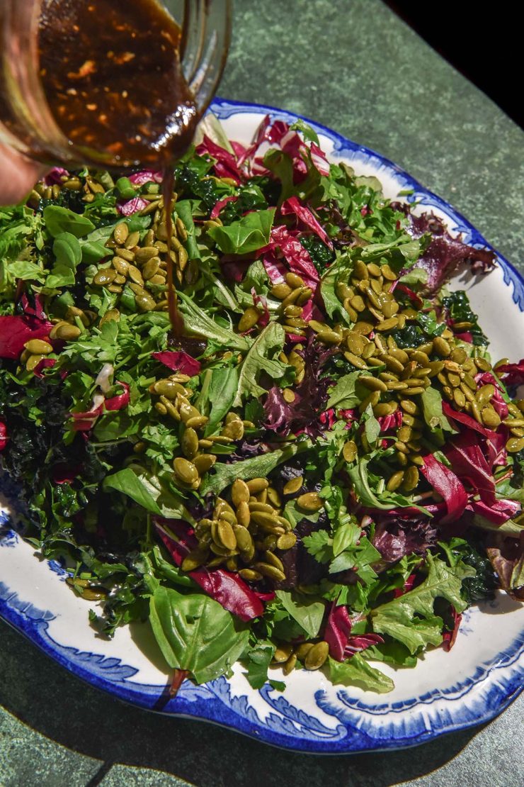 A side on image of low histamine salad dressing being poured onto a low histamine salad. The salad sits on a scalloped white and blue plate atop an olive green backdrop