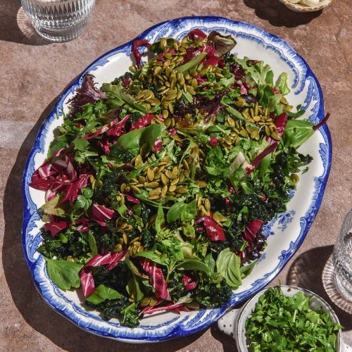 An aerial image of a low histamine salad on a white and blue scalloped serving dish. The salad sits atop a light brown backdrop and is surrounded by small bowls of salad ingredients and glasses of water.