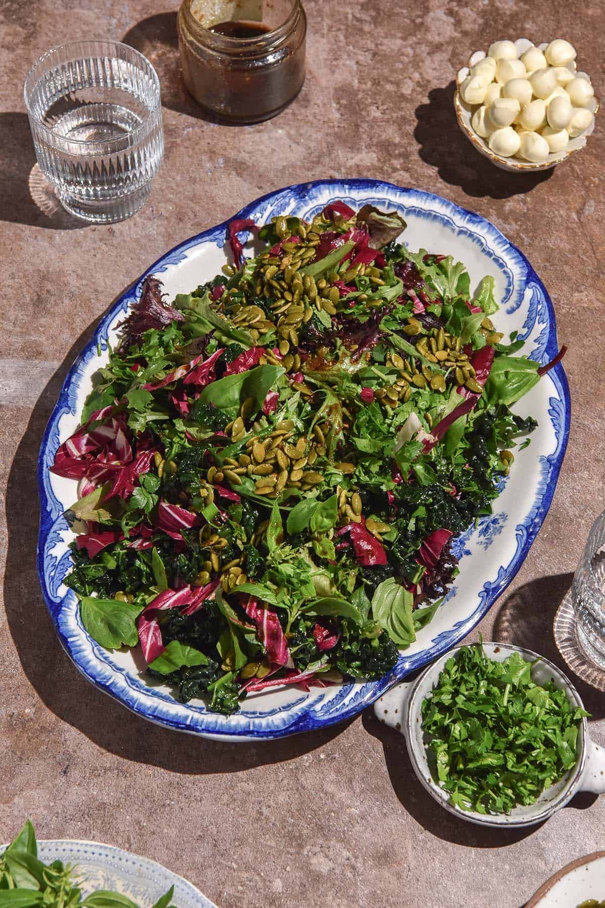 An aerial image of a low histamine salad on a white and blue scalloped serving platter. The platter sits on a brown backdrop and is surrounded by small white bowls of extra ingredients as well as water glasses