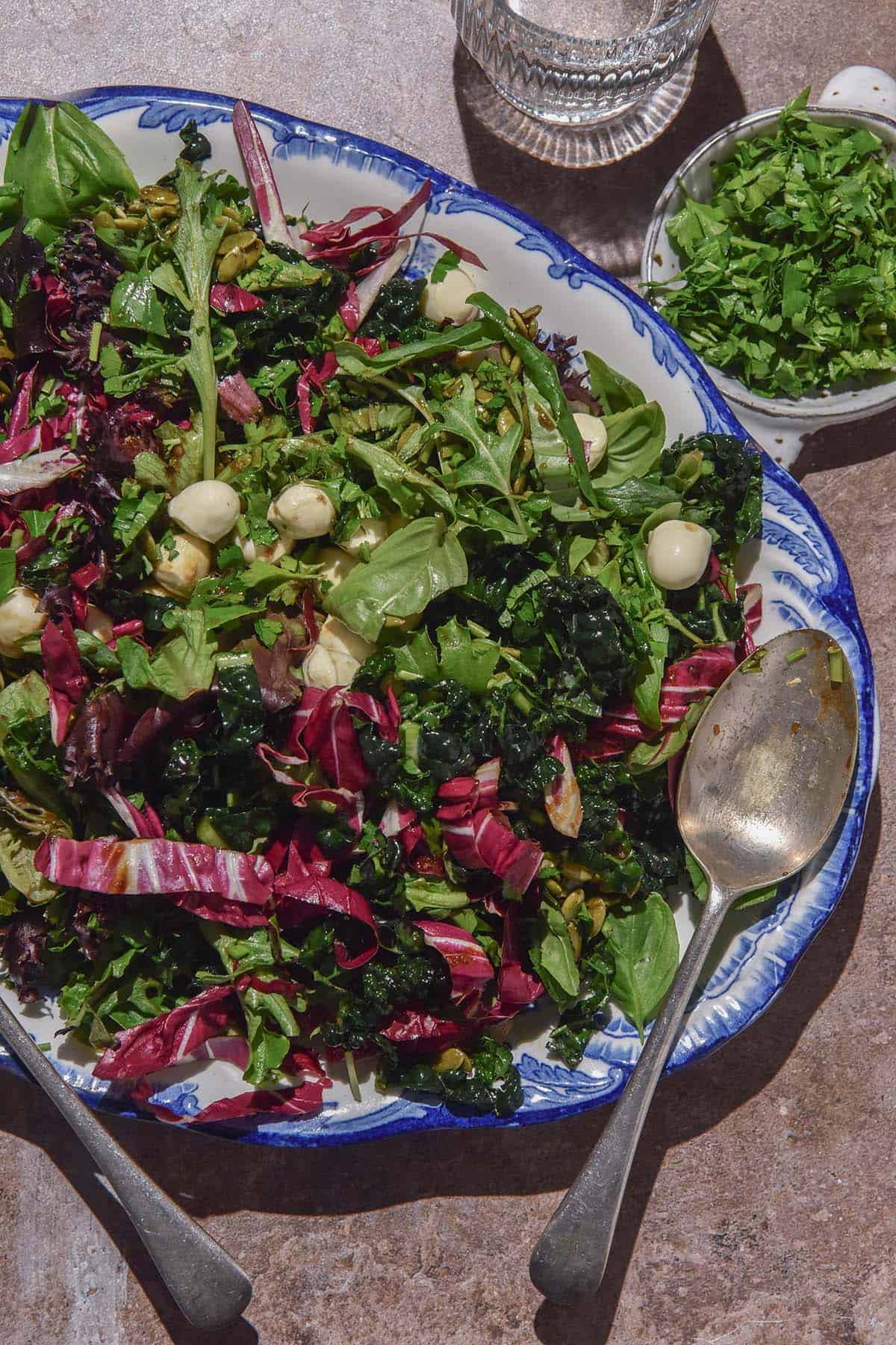 An aerial image of a low histamine salad on a blue and white scalloped plate atop a light brown backdrop. A sunlit glass of water and a white ceramic bowl filled with herbs sit to the right of the salad dish.
