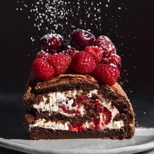 A side on image of a chocolate berry roulade on a white table against a dark backdrop. The roulade has been sliced to reveal the berries and cream inside. Icing sugar falls from the top of the image onto the berries atop the roulade