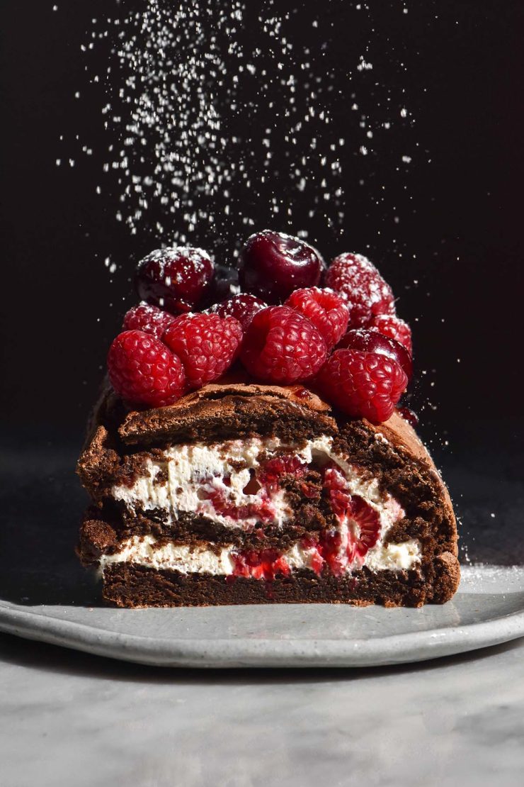 A side on image of a chocolate berry roulade on a white table against a dark backdrop. The roulade has been sliced to reveal the berries and cream inside. Icing sugar falls from the top of the image onto the berries atop the roulade