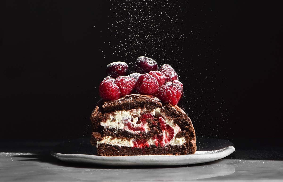 A moody image of a gluten free chocolate roulade filled with cream and topped with berries. Icing sugar sprinkles down onto the roulade which is set against a dark backdrop