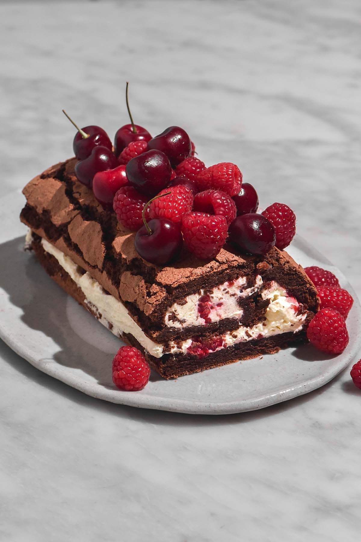 A side on image of a chocolate berry roulade covered in extra berries on a white ceramic plate atop a white marble table