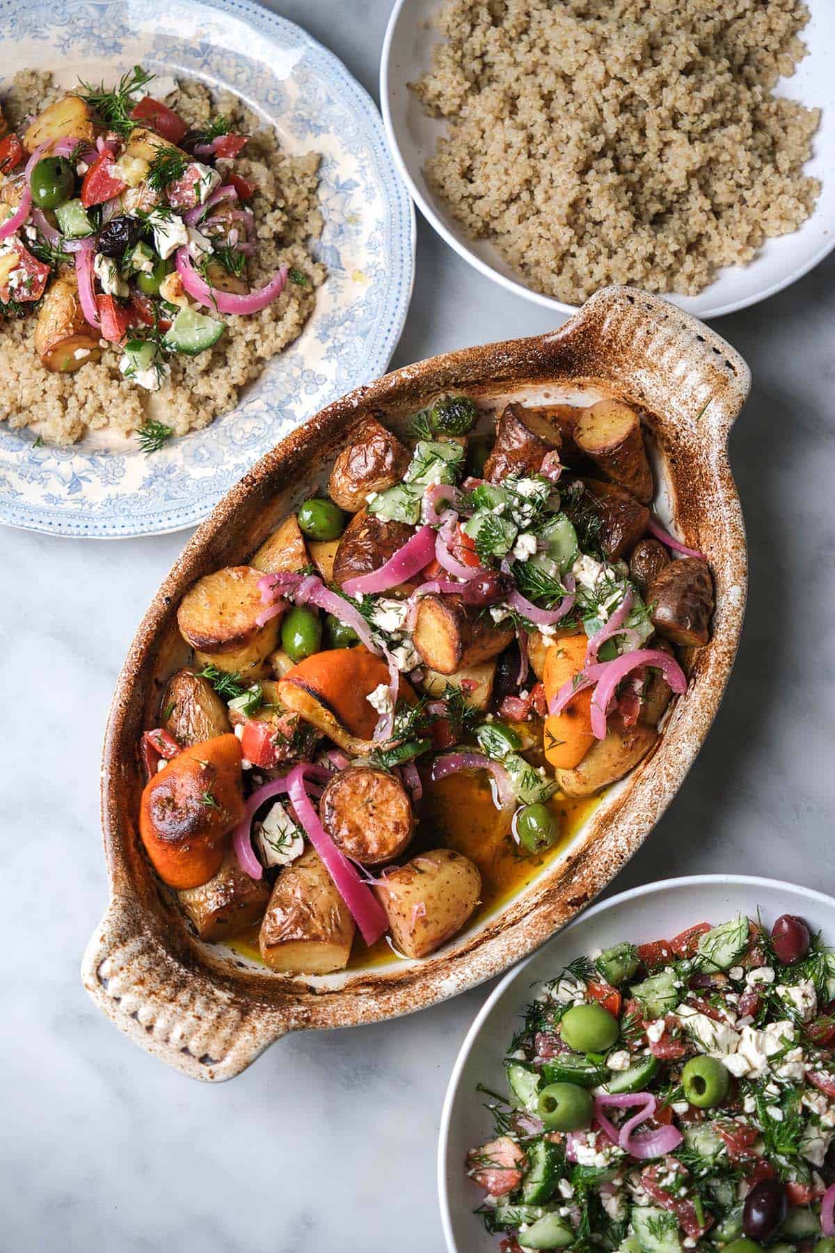 An aerial image of a white ceramic dish filled with Greek salad potatoes on a white marble table. The dish is surrounded by white bowls filled with Greek salad and cooked quinoa, and a plate of the final dish sits to the top left of the potatoes