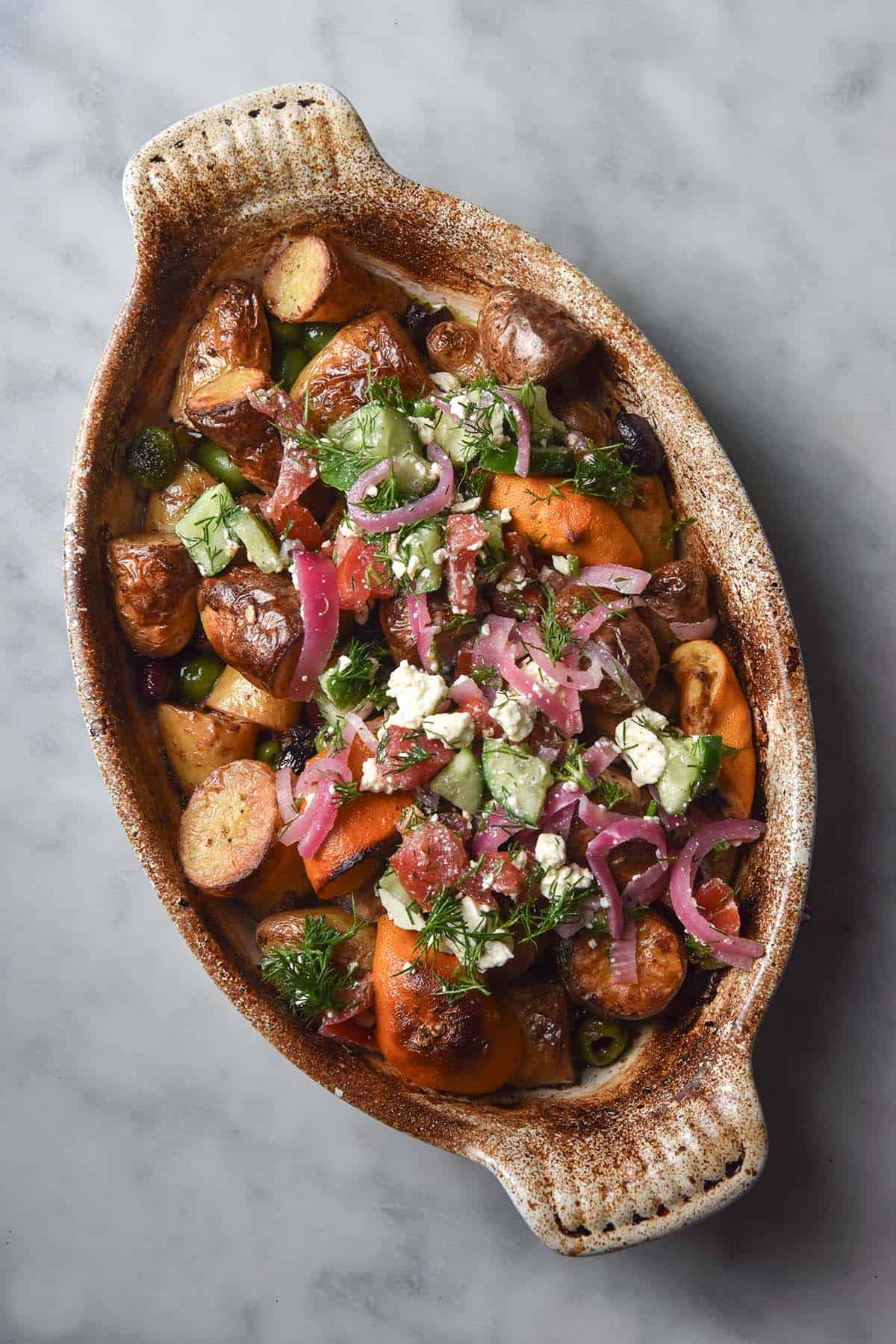 An aerial image of a white ceramic baking dish filled with Greek salad potatoes atop a white marble table