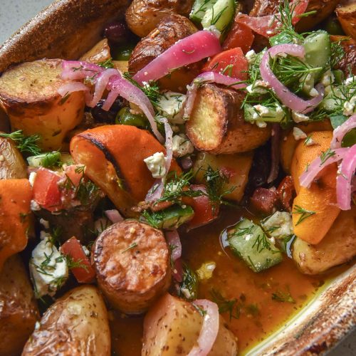 A close up image of a ceramic baking dish filled with Greek salad potatoes. The dish sits atop a white speckled stone bench top.