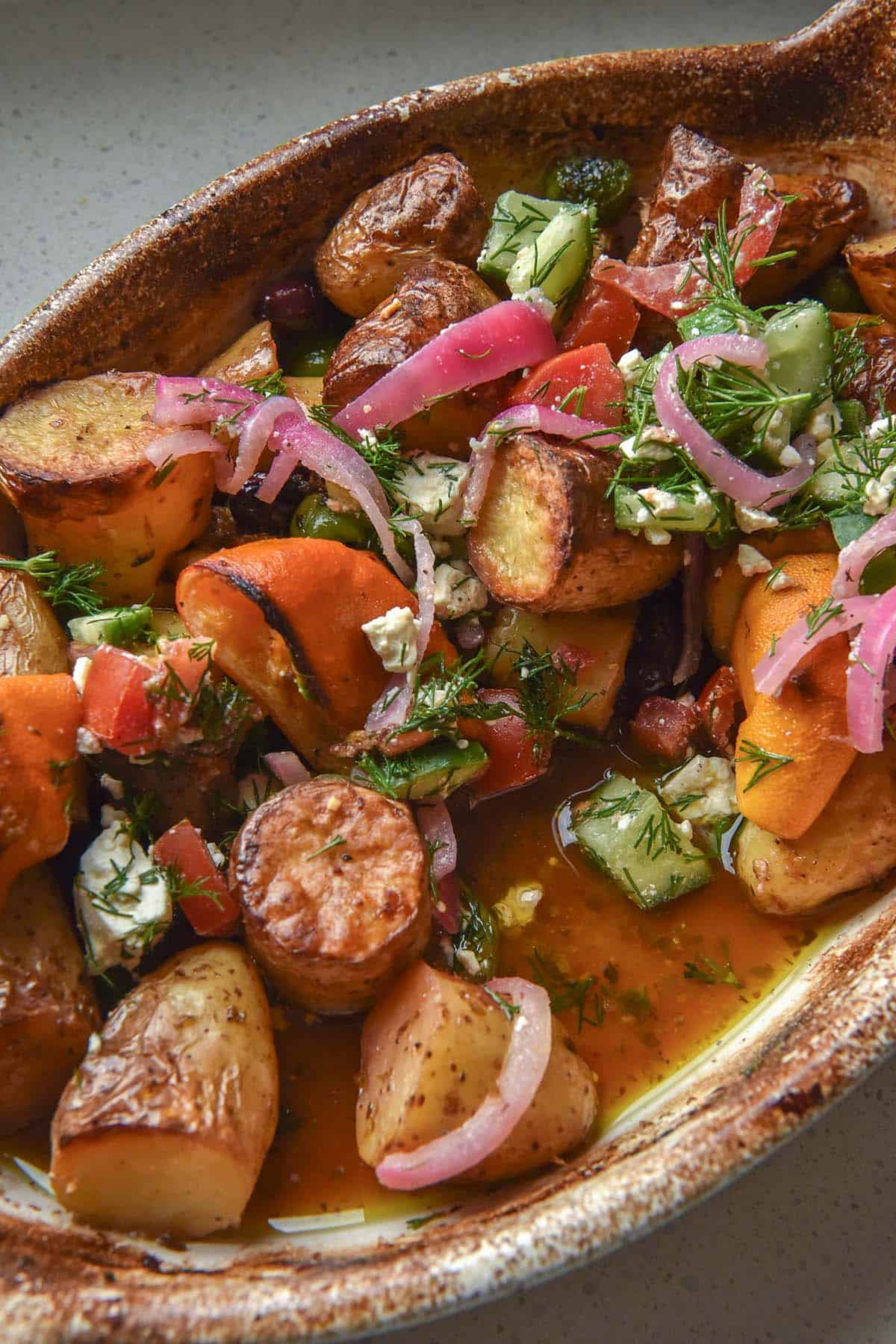 A close up image of a ceramic baking dish filled with Greek salad potatoes. The dish sits atop a white speckled stone bench top.