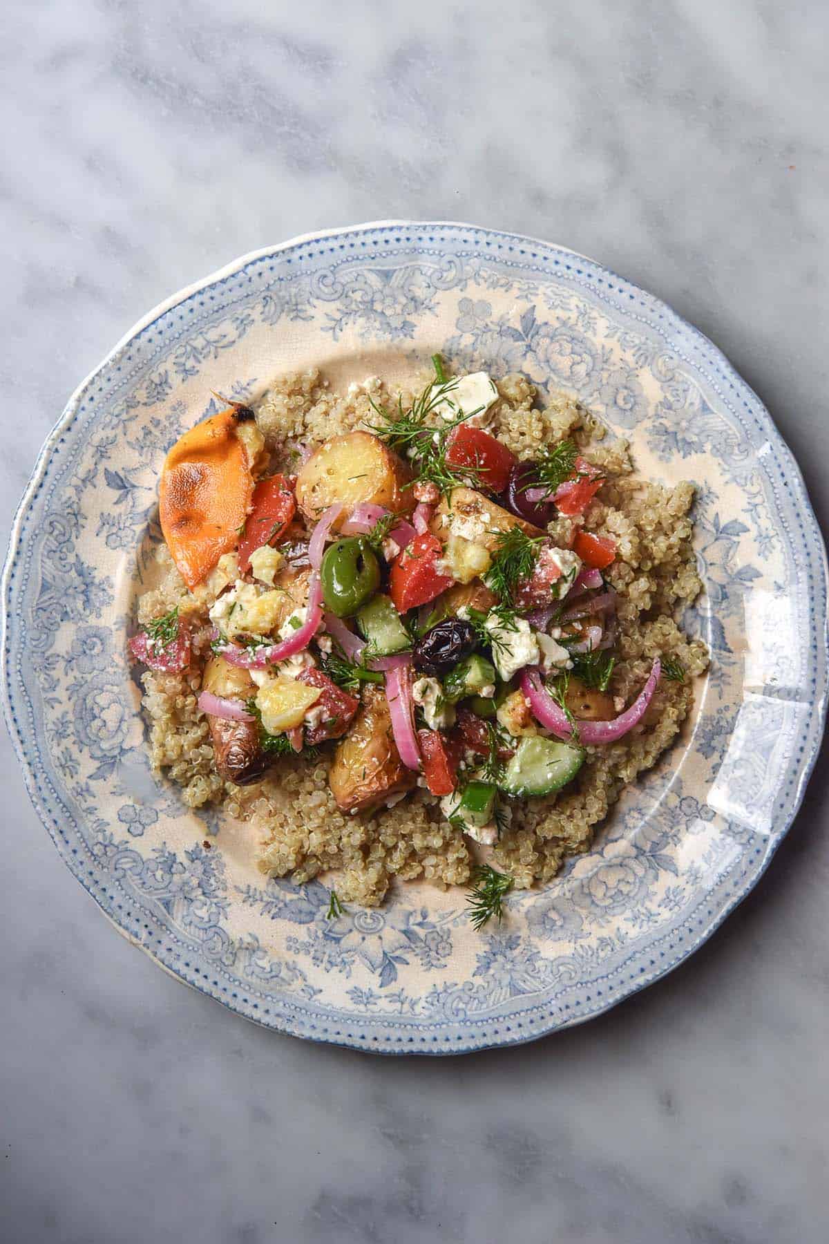 An aerial image of a blue and white patterned plate topped with quinoa and Greek salad potatoes. The dish sits atop a white marble table.