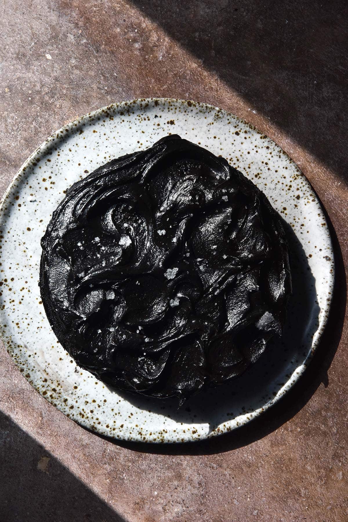 An aerial image of a cake topped with black buttercream on a white speckled ceramic plate atop a brown backdrop in contrasting sunlight