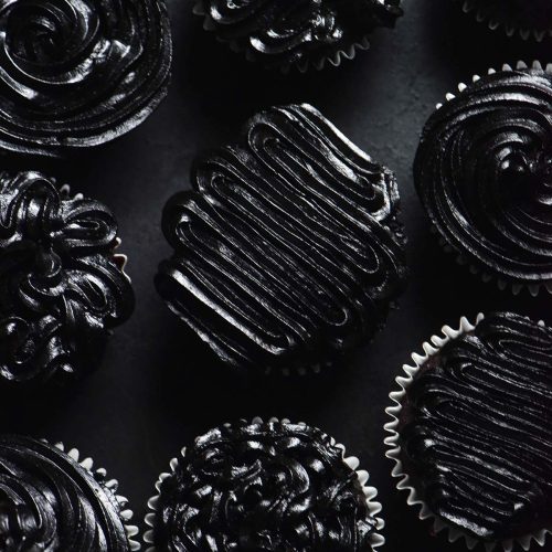 An aerial image of cupcakes topped with black buttercream in different patterns on a black backdrop