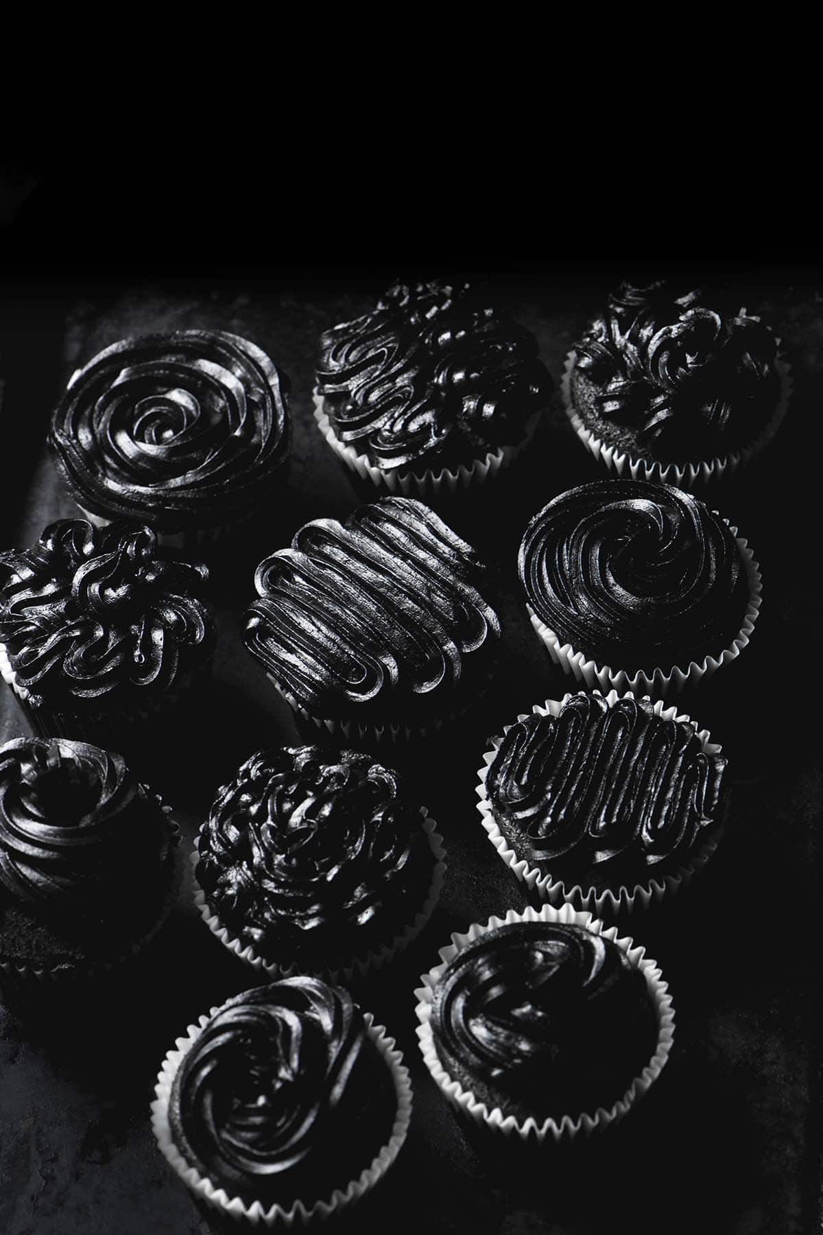 A dark and moody image of cupcakes topped with blacking buttercream in different patterns on a black surface against a black backdrop