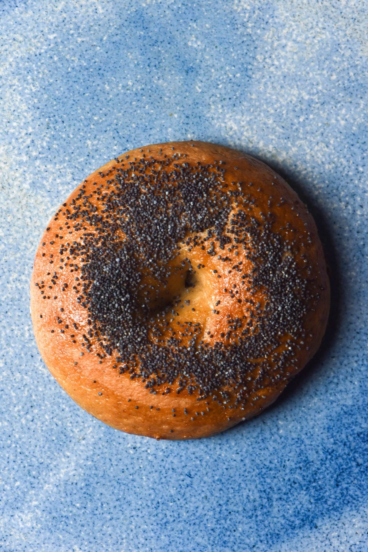 An aerial image of a gluten free sourdough bagel topped with poppyseeds on a bright blue ceramic plate