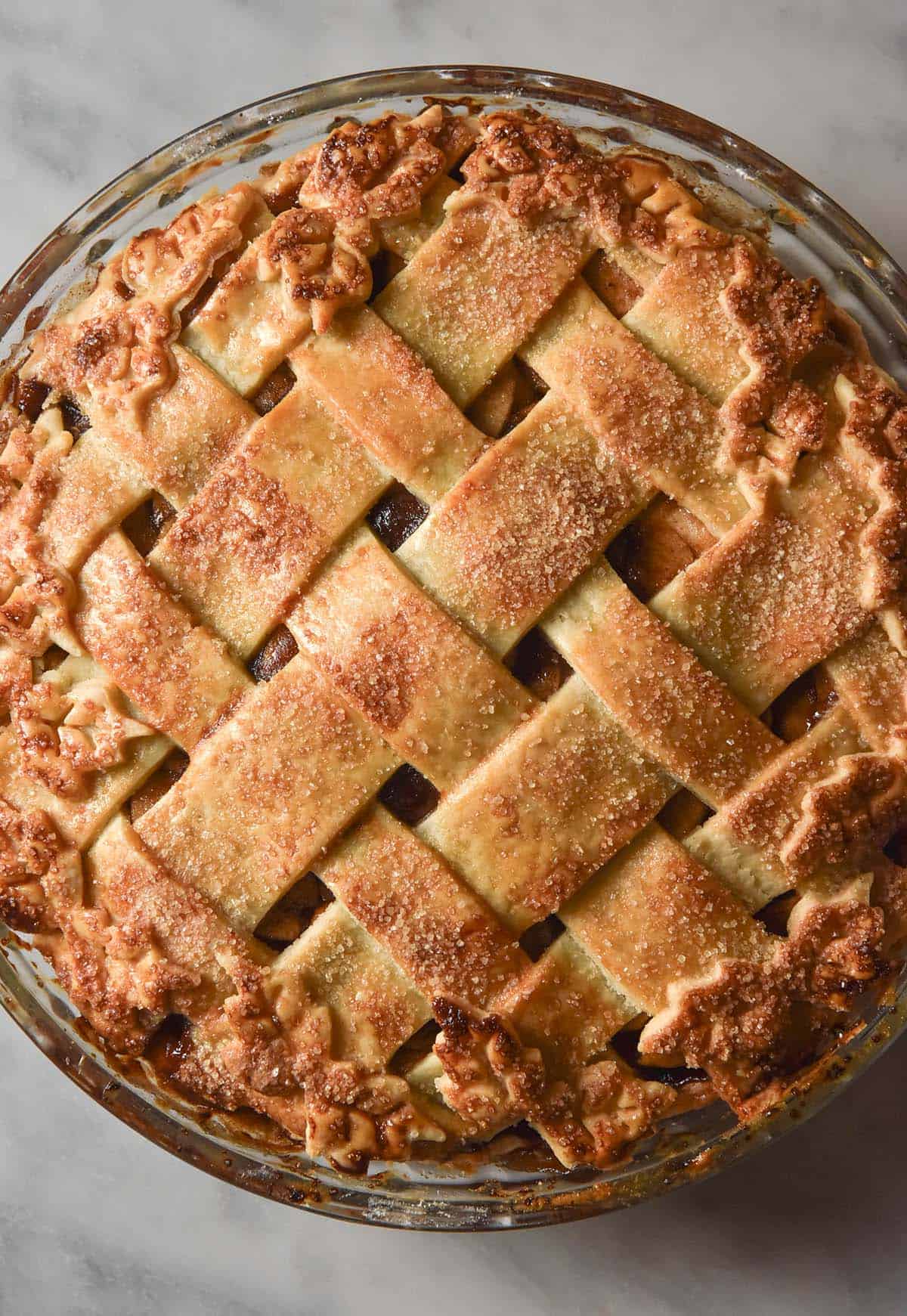 An aerial image of a golden brown gluten free apple pie with a latticework top surrounded by pastry leaves. The pie sits on a white marble table