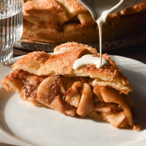A brightly lit side on image of a slice of gluten free apple pie on a white place. A hand extends down from the top of the image to drizzle cream onto the apple pie and the remaining pie sits in the background