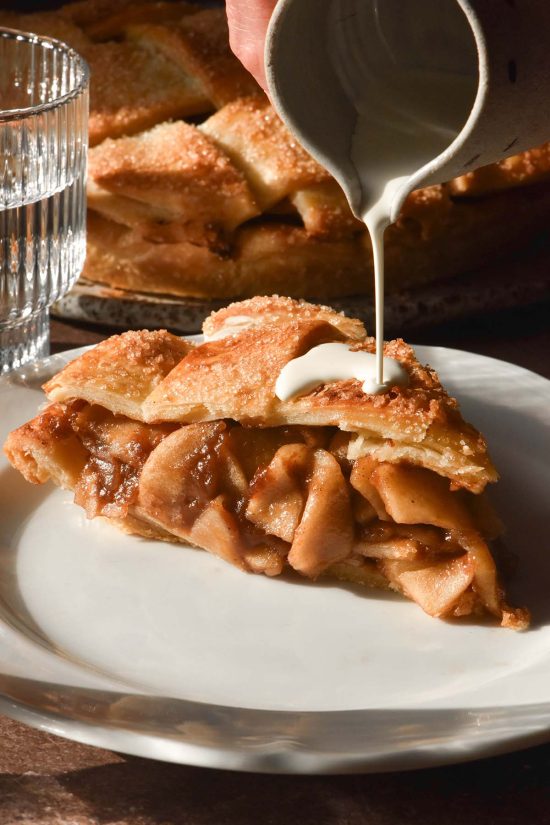 A brightly lit side on image of a slice of gluten free apple pie on a white place. A hand extends down from the top of the image to drizzle cream onto the apple pie and the remaining pie sits in the background