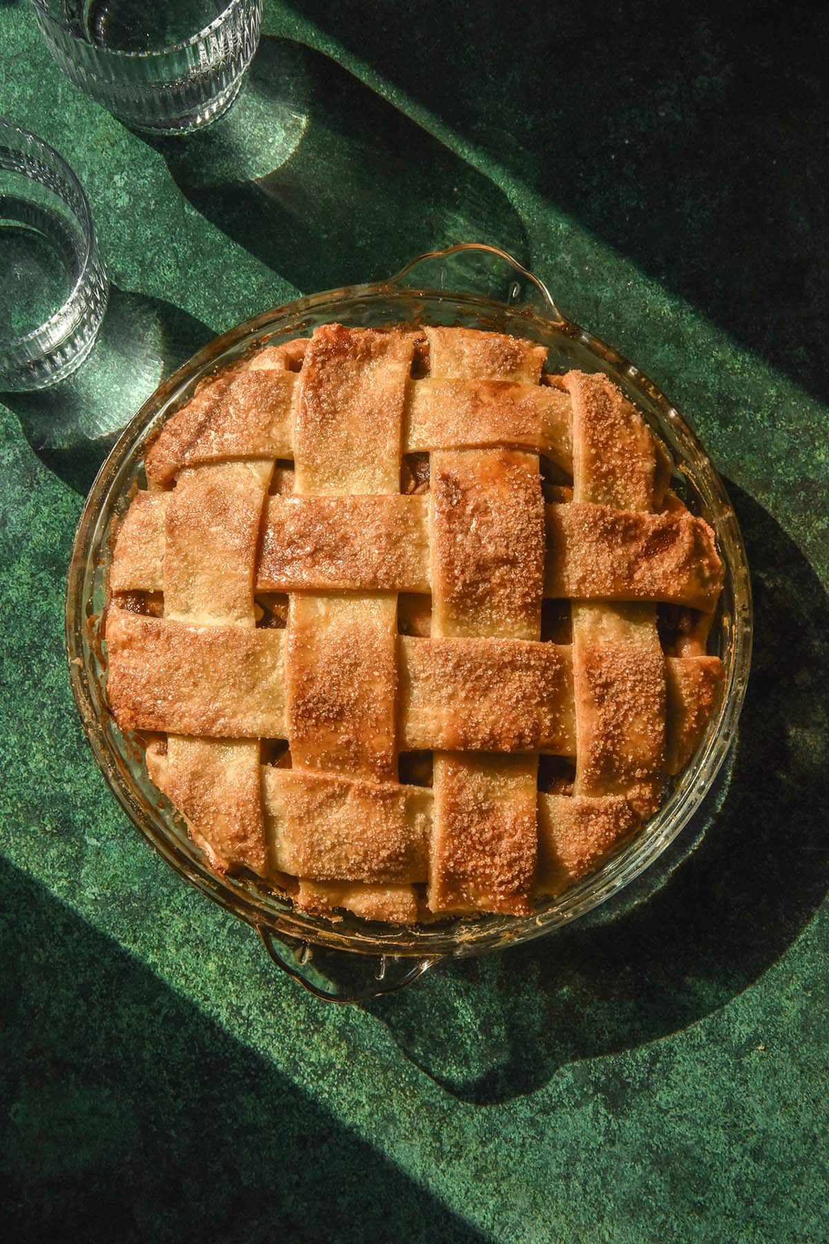 An aerial image of a gluten free apple pie in contrasting sunlight. The pie sits atop a dark green backdrop and sunlit water glasses sit to the top left of the image