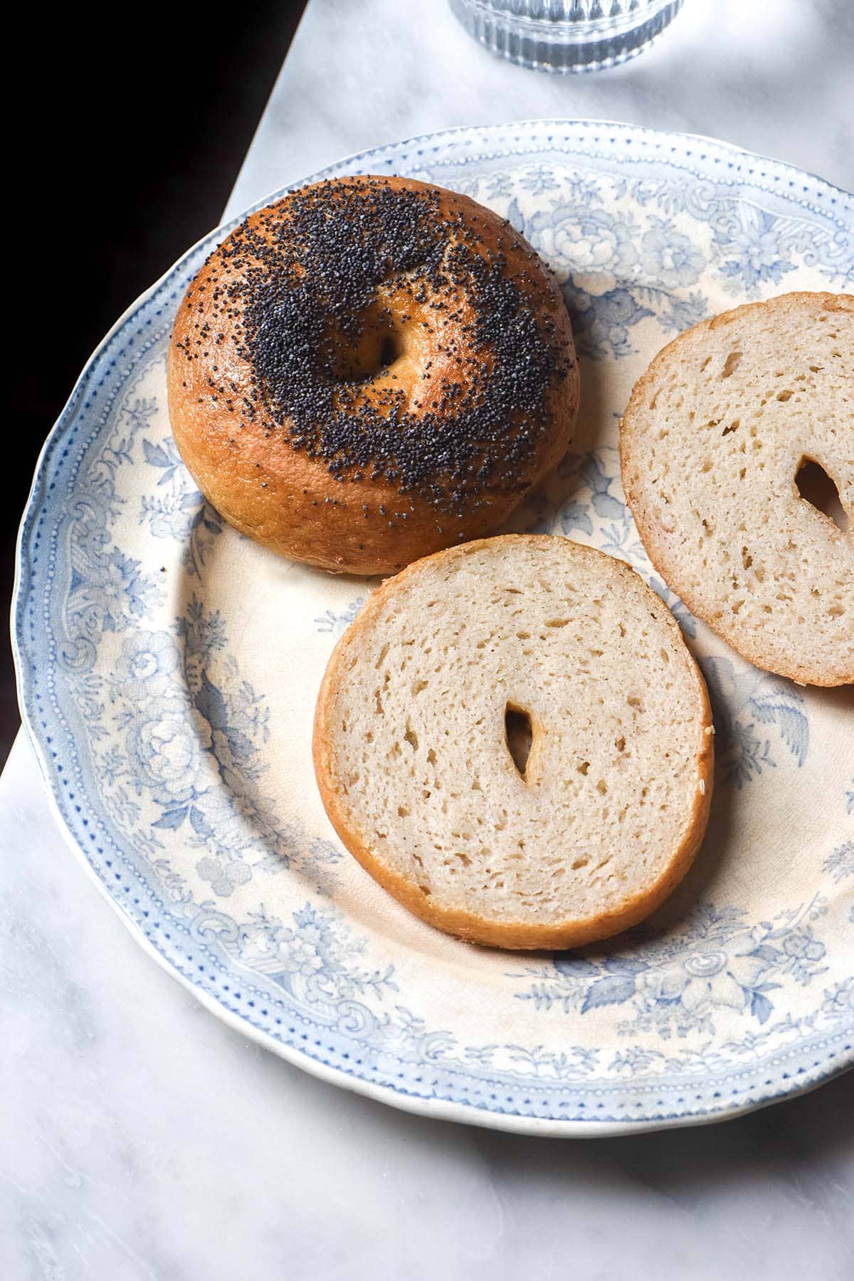 An aerial image of a gluten free sourdough poppyseed bagel alongside a halved bagel on a blue and white plate atop a white marble table