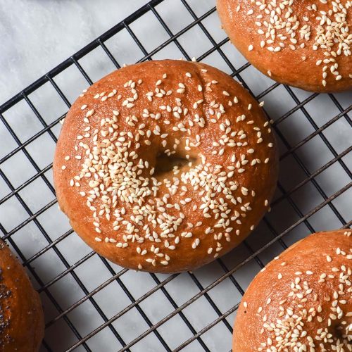 An aerial image of gluten free sourdough bagels topped with sesame seeds on a wire cooling rack atop a white marble table