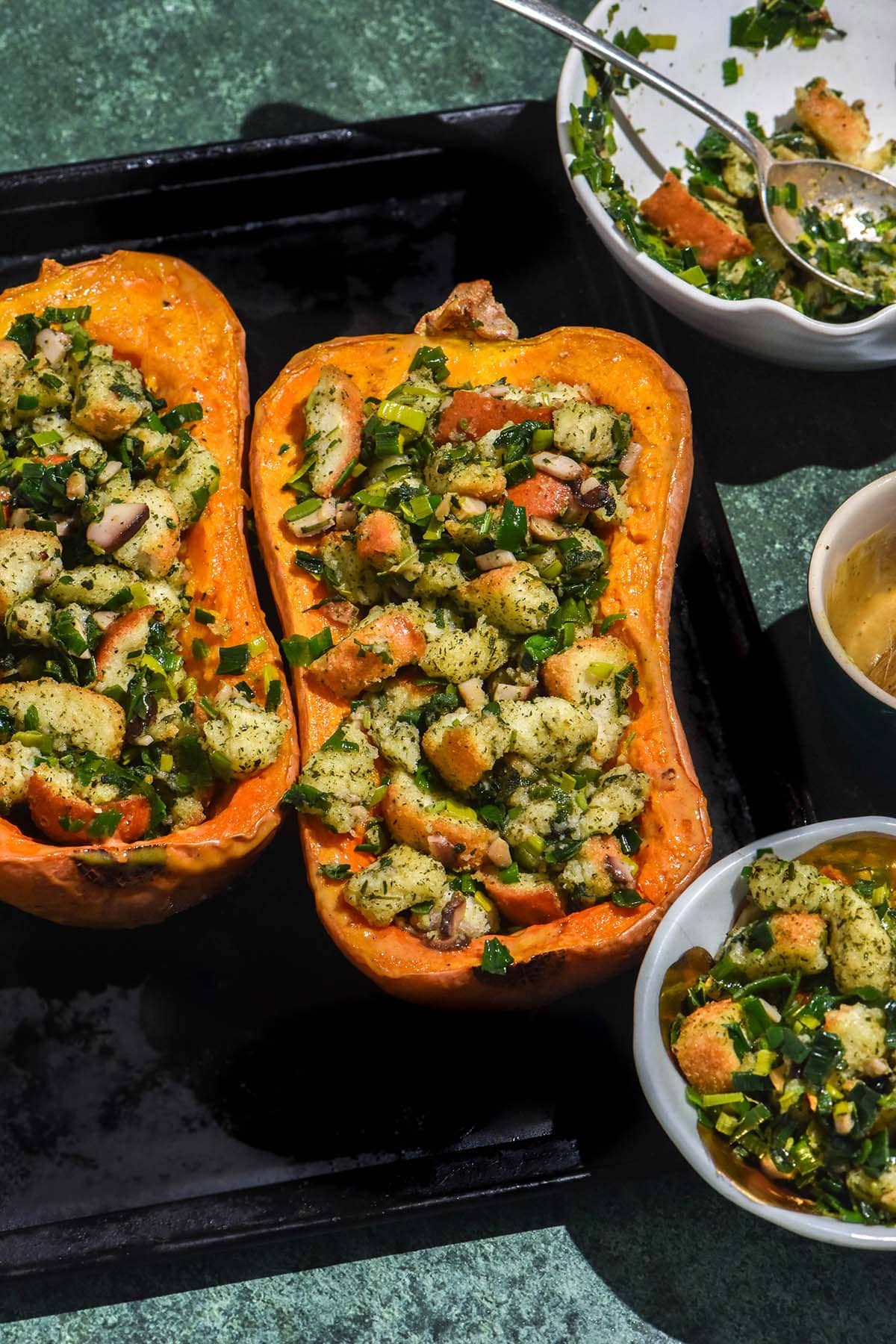 An aerial image of stuffing filled pumpkins on a black baking tray atop a green backdrop. The pumpkins are surrounded by small bowls of extra stuffing and glaze