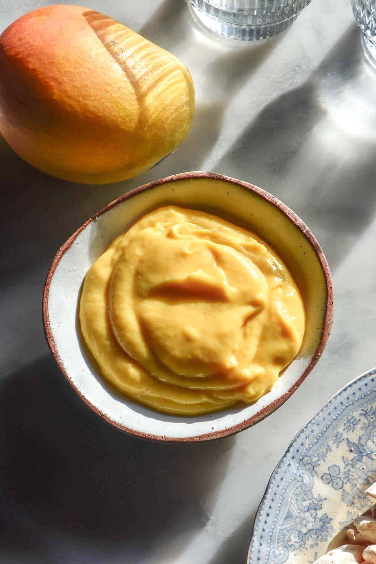 A sunlit aerial image of a bowl of mango curd on a white marble table. A mango sits in the top left corner and a plate of mango curd Eton mess sits in the bottom right corner. Two sunlit glasses of water sit in the top right corner of the image.
