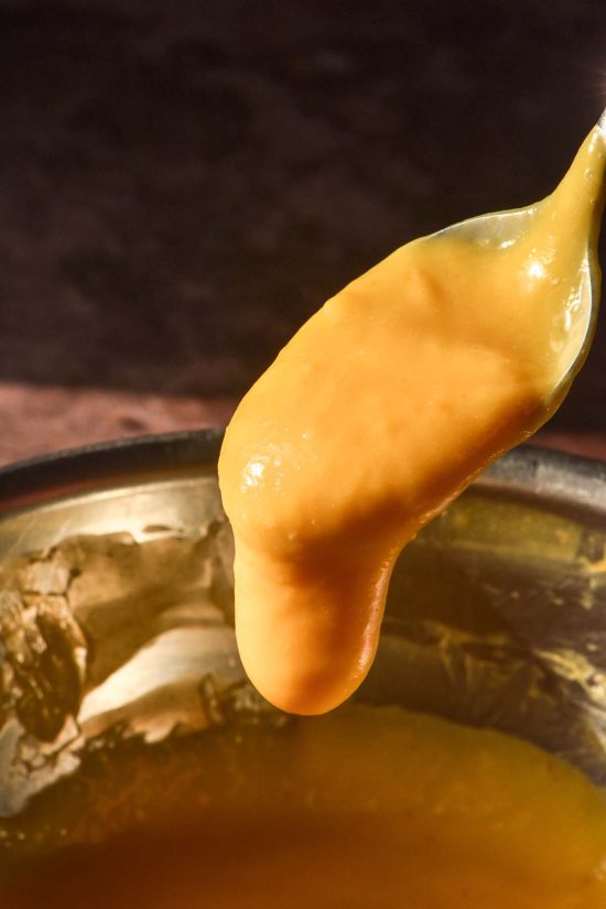 A moody side on image of a spoon topped with dairy free mango curd against a dark backdrop. The curd is dripping back down into the bowl below the spoon.