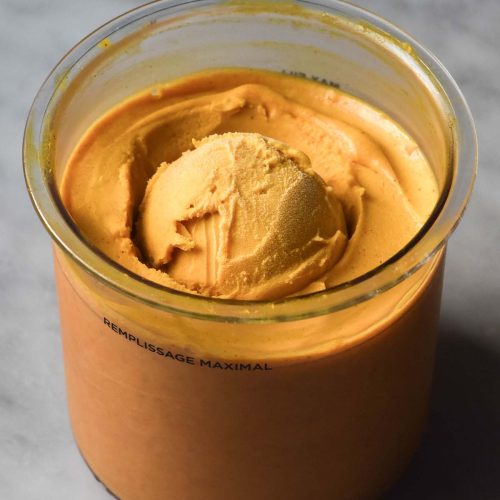 A side on image of a tub of pumpkin Ninja Creami ice cream on a white marble table. The ice cream has a scoop of ice cream placed in the centre of the tub.