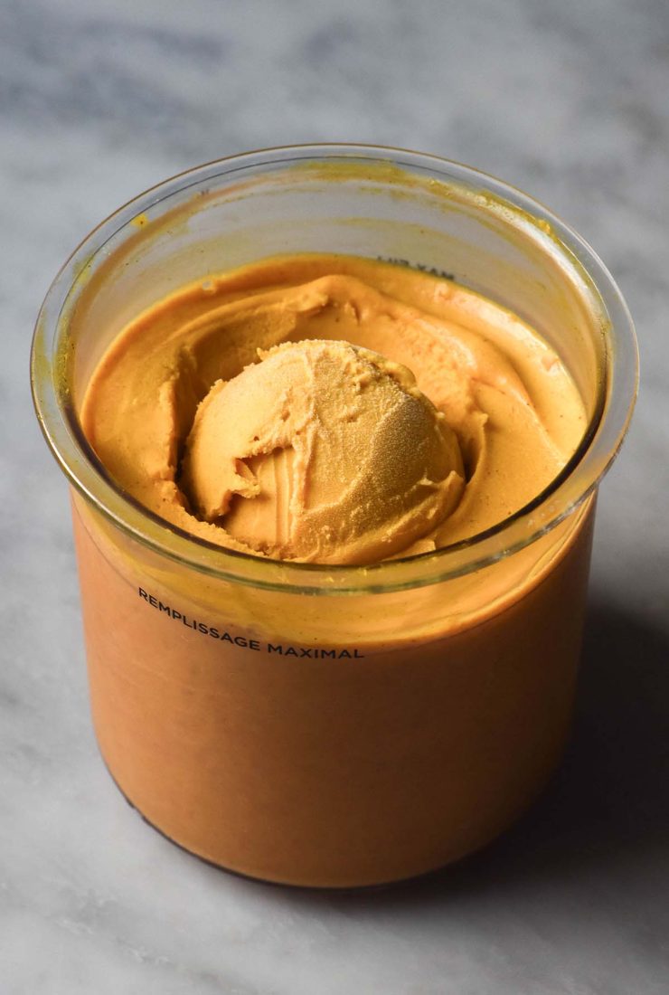 A side on image of a tub of pumpkin Ninja Creami ice cream on a white marble table. The ice cream has a scoop of ice cream placed in the centre of the tub.