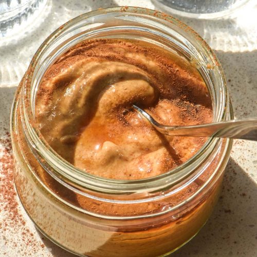 A sunlit aerial image of a blended pumpkin spice chia pudding in a jar on a white stone bench top. The pudding has been drizzled with maple syrup and sprinkled with extra pumpkin spice. Two sunlit water glasses sit to either side of the pudding