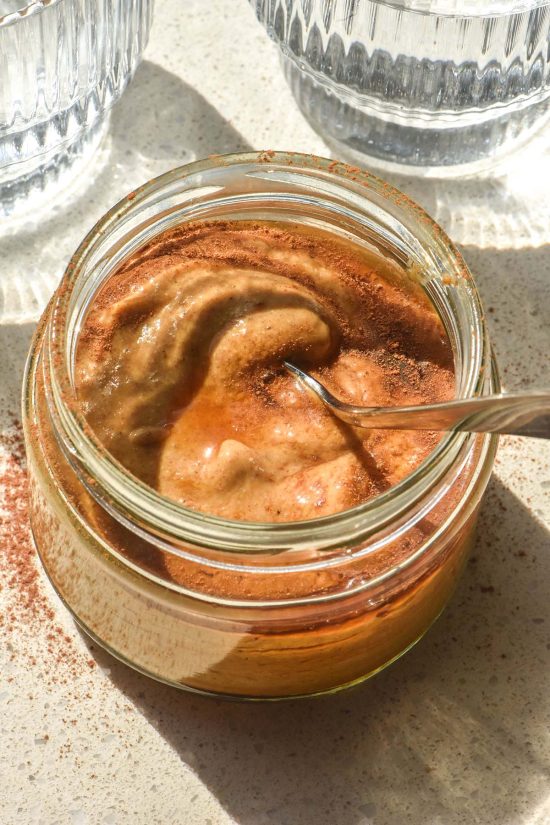 A sunlit aerial image of a blended pumpkin spice chia pudding in a jar on a white stone bench top. The pudding has been drizzled with maple syrup and sprinkled with extra pumpkin spice. Two sunlit water glasses sit to either side of the pudding