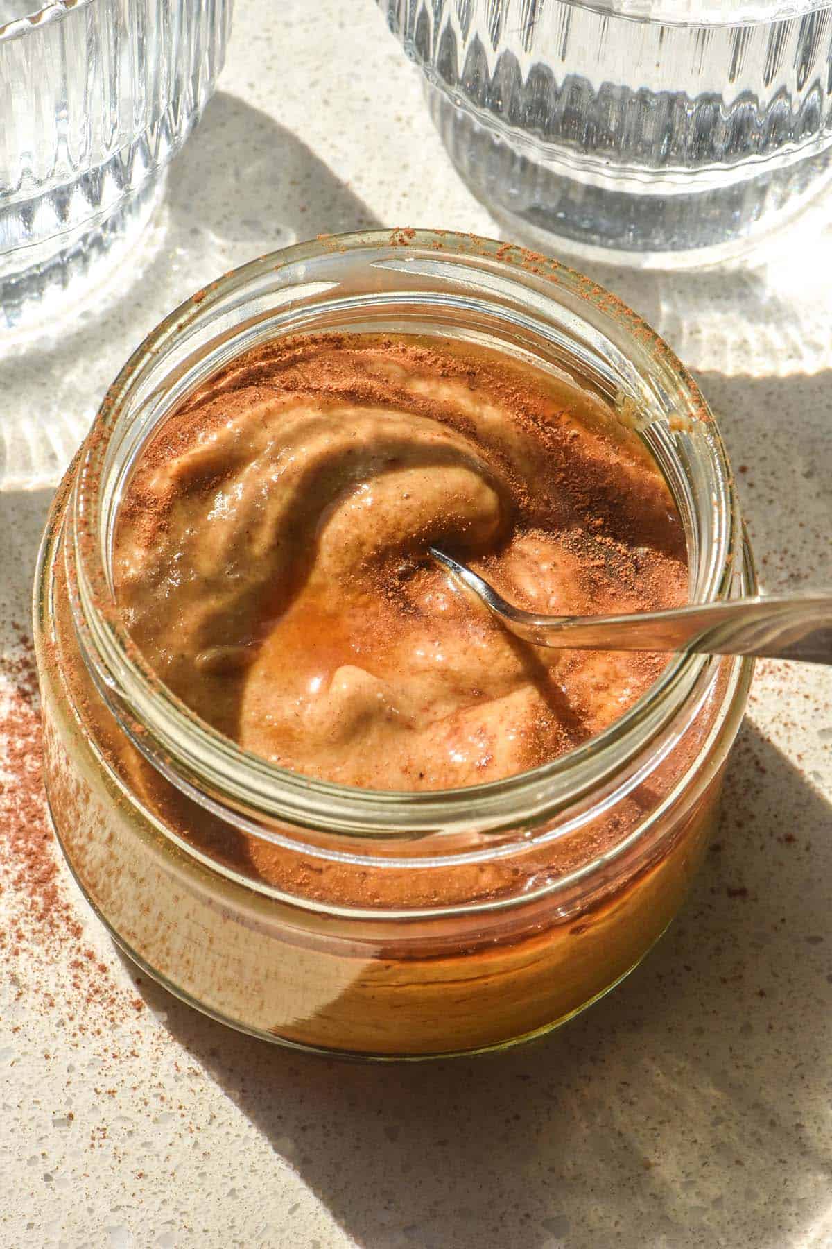 A sunlit aerial image of a blended pumpkin spice chia pudding in a jar on a white stone bench top. The pudding has been drizzled with maple syrup and sprinkled with extra pumpkin spice. Two sunlit water glasses sit to either side of the pudding