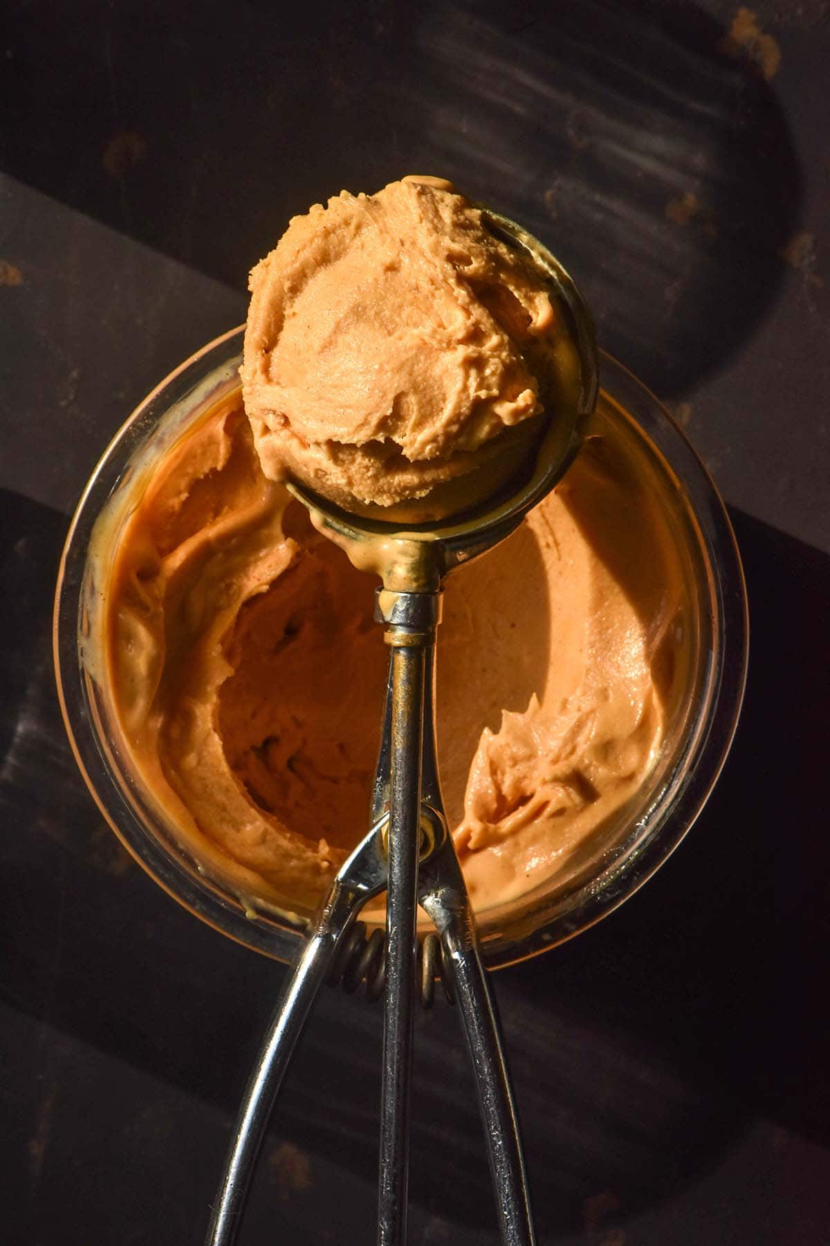 A moody image of a tub of sweet potato Ninja Creami ice cream on a black backdrop in contrasting sunlight. An ice cream scoop with a scoop of the ice cream rests on top of the tub