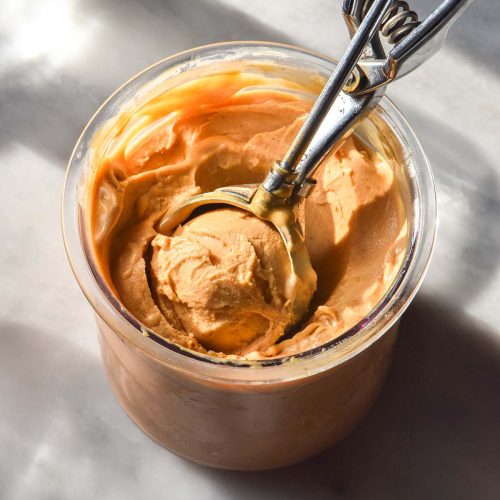 An image of a tub of sweet potato Ninja Creami ice cream on a white marble table in contrasting sunlight. A scoop sits in the tub filled with the ice cream
