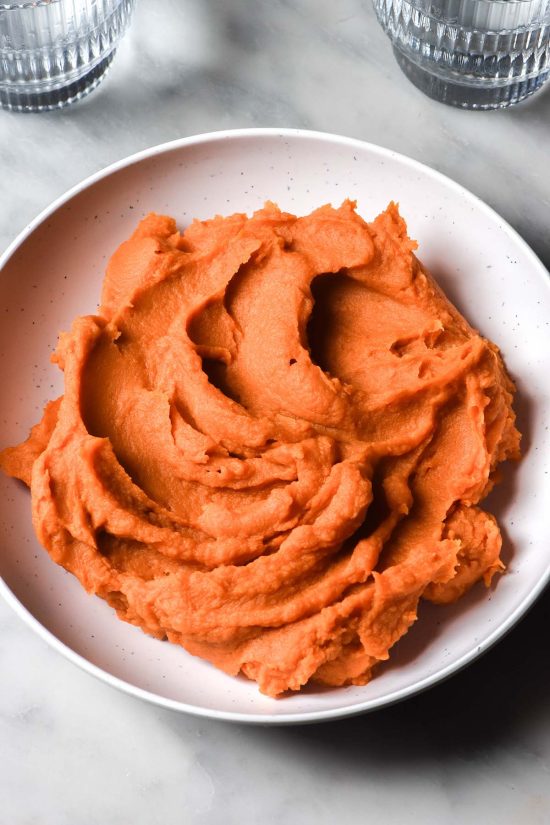 An aerial image of a white bowl filled with sweet potato puree atop a white marble table. Two glasses of water sit on either side of the bowl in the top corners of the image.